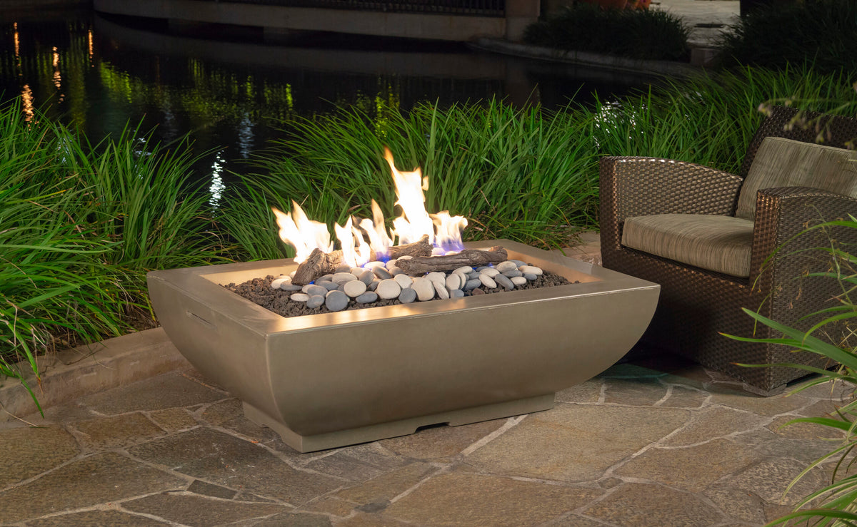 American Fyre Design Bordeaux Rectangle Fire Bowl lit on a stone patio beside a wicker lounge chair