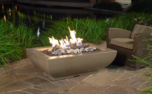 American Fyre Design Bordeaux Rectangle Fire Bowl lit on a stone patio beside a wicker lounge chair