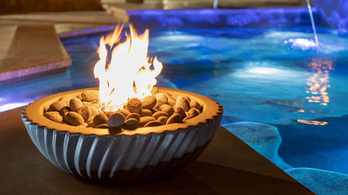 The Outdoor Plus Sedona fire bowl burning at the edge of a lit swimming pool