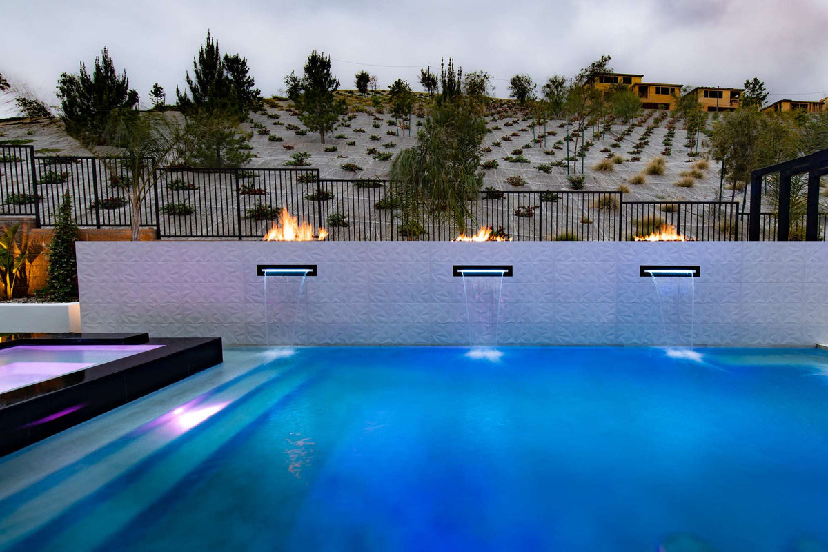 A custom The Outdoor Plus fire feature burning beside a lit pool at night