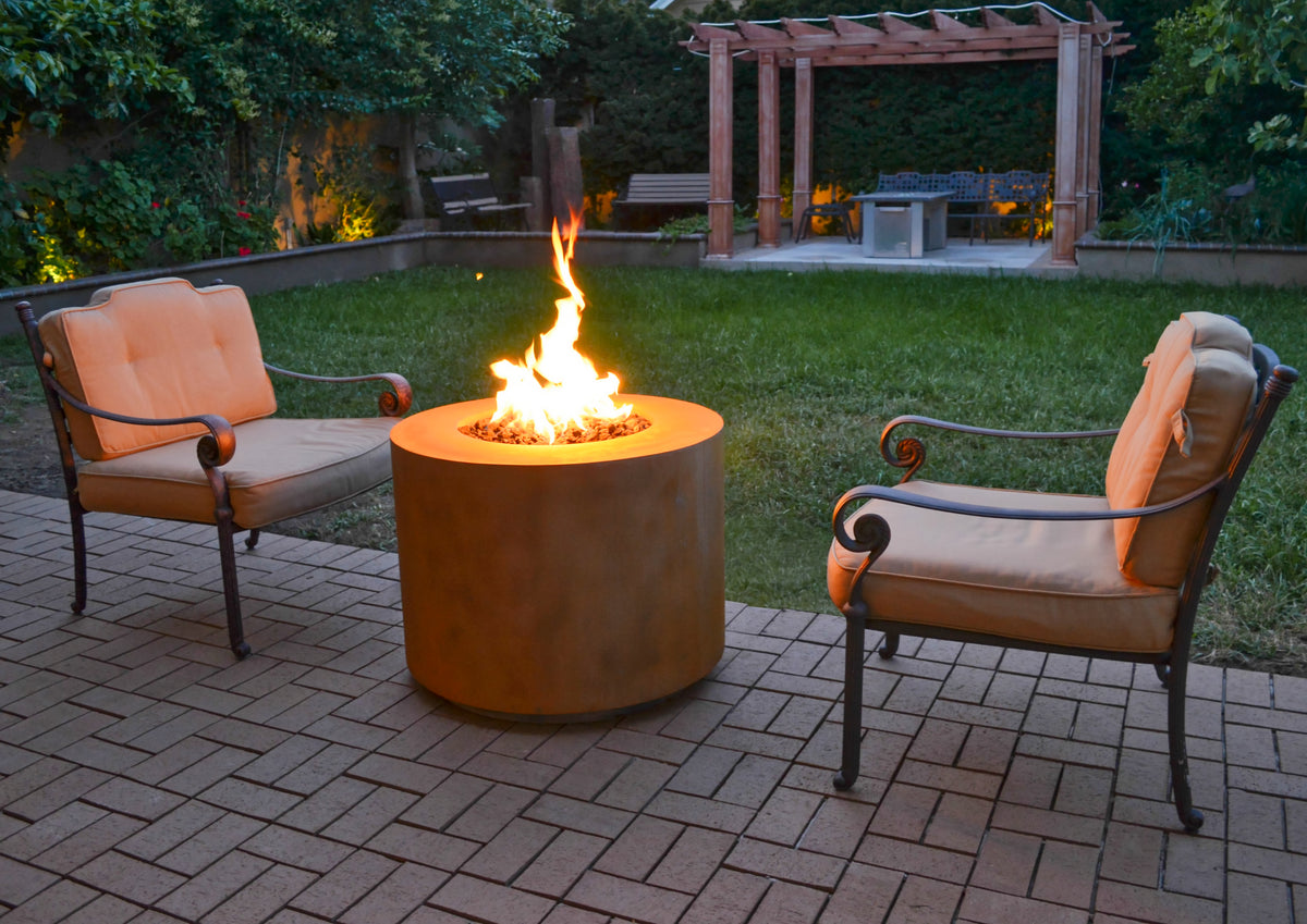 The Outdoor Plus corten steel fire pit lit between two lounge chairs on a patio